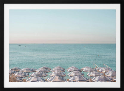 A high-angle photograph of a beach packed with blue and white striped umbrellas, with the turquoise ocean stretching out to a pale blue sky in the background. The entire image is enclosed in a silver frame. Poster