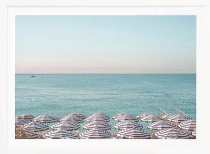 A high-angle photograph of a beach packed with blue and white striped umbrellas, with the turquoise ocean stretching out to a pale blue sky in the background. The entire image is enclosed in a silver frame. Poster