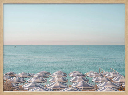 A high-angle photograph of a beach packed with blue and white striped umbrellas, with the turquoise ocean stretching out to a pale blue sky in the background. The entire image is enclosed in a silver frame. Poster