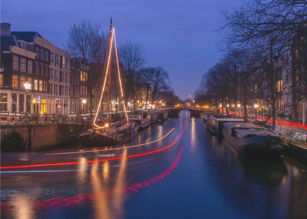 A long-exposure photograph of an Amsterdam canal at dusk. The scene is bathed in a deep blue light, with warm yellow and orange lights from street lamps and buildings reflecting on the water. A sailboat is decorated with glowing string lights, and red and white light trails from a passing boat create streaks across the canal. Houseboats are moored along the sides, and a bridge is visible in the distance. Poster