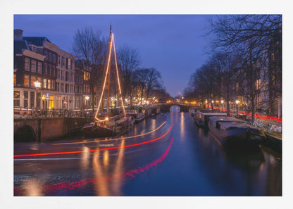 A long-exposure photograph of an Amsterdam canal at dusk. The scene is bathed in a deep blue light, with warm yellow and orange lights from street lamps and buildings reflecting on the water. A sailboat is decorated with glowing string lights, and red and white light trails from a passing boat create streaks across the canal. Houseboats are moored along the sides, and a bridge is visible in the distance. Poster