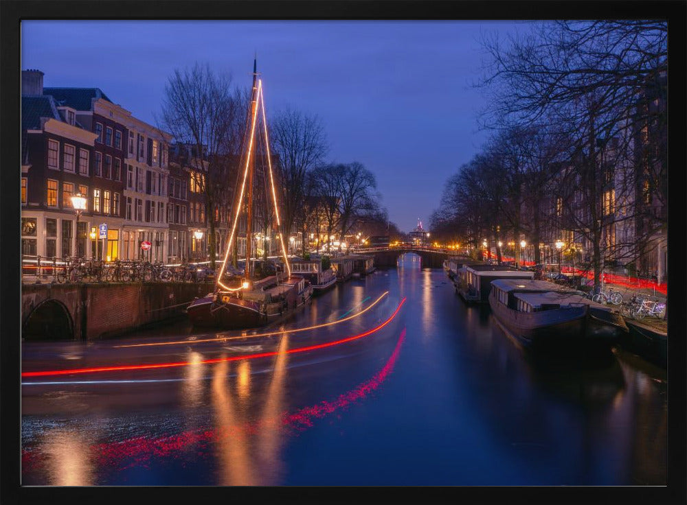 A long-exposure photograph of an Amsterdam canal at dusk. The scene is bathed in a deep blue light, with warm yellow and orange lights from street lamps and buildings reflecting on the water. A sailboat is decorated with glowing string lights, and red and white light trails from a passing boat create streaks across the canal. Houseboats are moored along the sides, and a bridge is visible in the distance. Poster