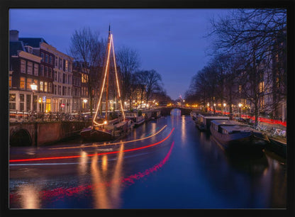 A long-exposure photograph of an Amsterdam canal at dusk. The scene is bathed in a deep blue light, with warm yellow and orange lights from street lamps and buildings reflecting on the water. A sailboat is decorated with glowing string lights, and red and white light trails from a passing boat create streaks across the canal. Houseboats are moored along the sides, and a bridge is visible in the distance. Poster