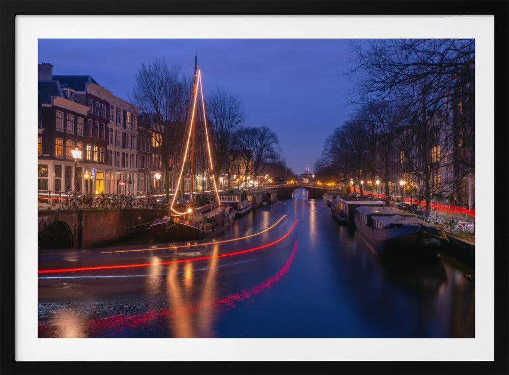 A long-exposure photograph of an Amsterdam canal at dusk. The scene is bathed in a deep blue light, with warm yellow and orange lights from street lamps and buildings reflecting on the water. A sailboat is decorated with glowing string lights, and red and white light trails from a passing boat create streaks across the canal. Houseboats are moored along the sides, and a bridge is visible in the distance. Poster