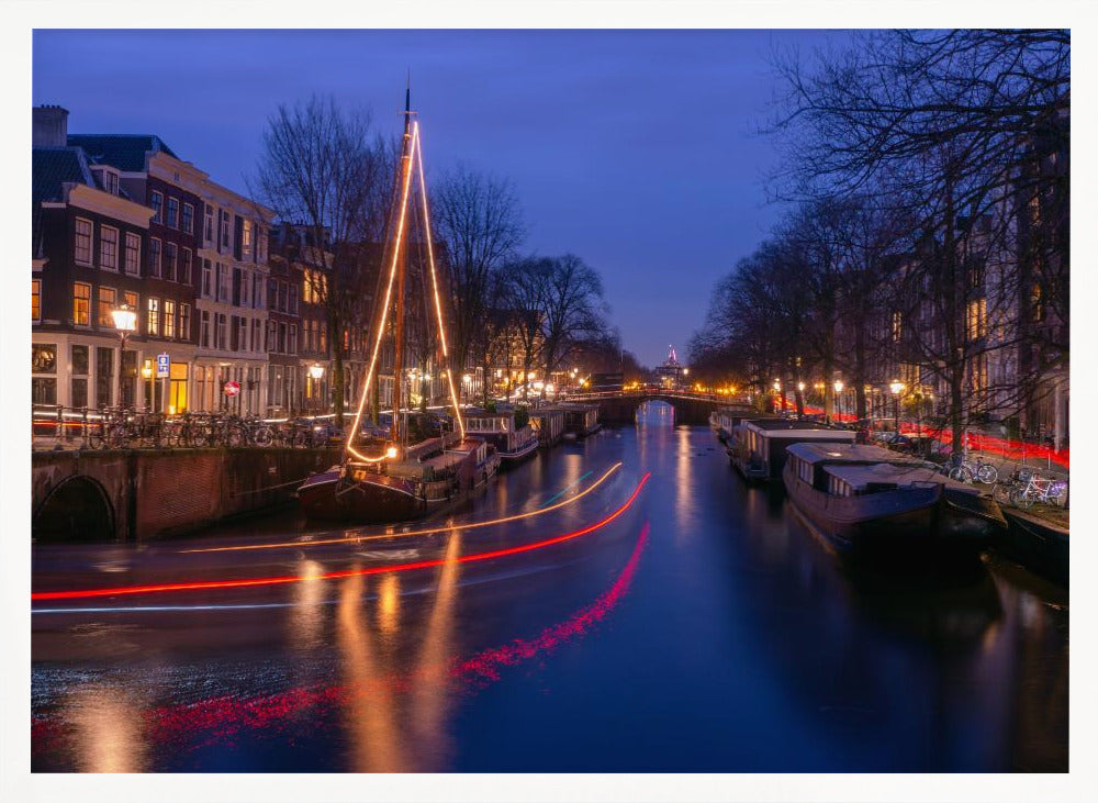 A long-exposure photograph of an Amsterdam canal at dusk. The scene is bathed in a deep blue light, with warm yellow and orange lights from street lamps and buildings reflecting on the water. A sailboat is decorated with glowing string lights, and red and white light trails from a passing boat create streaks across the canal. Houseboats are moored along the sides, and a bridge is visible in the distance. Poster