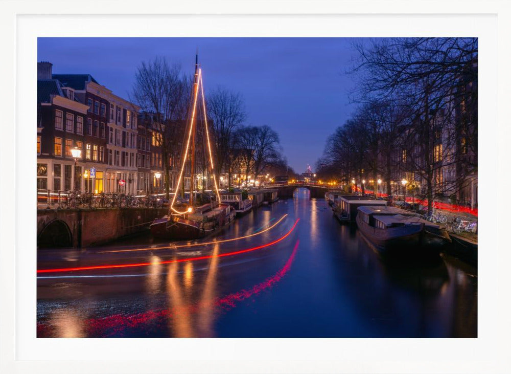 A long-exposure photograph of an Amsterdam canal at dusk. The scene is bathed in a deep blue light, with warm yellow and orange lights from street lamps and buildings reflecting on the water. A sailboat is decorated with glowing string lights, and red and white light trails from a passing boat create streaks across the canal. Houseboats are moored along the sides, and a bridge is visible in the distance. Poster