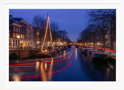 A long-exposure photograph of an Amsterdam canal at dusk. The scene is bathed in a deep blue light, with warm yellow and orange lights from street lamps and buildings reflecting on the water. A sailboat is decorated with glowing string lights, and red and white light trails from a passing boat create streaks across the canal. Houseboats are moored along the sides, and a bridge is visible in the distance. Poster