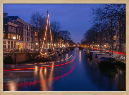 A long-exposure photograph of an Amsterdam canal at dusk. The scene is bathed in a deep blue light, with warm yellow and orange lights from street lamps and buildings reflecting on the water. A sailboat is decorated with glowing string lights, and red and white light trails from a passing boat create streaks across the canal. Houseboats are moored along the sides, and a bridge is visible in the distance. Poster