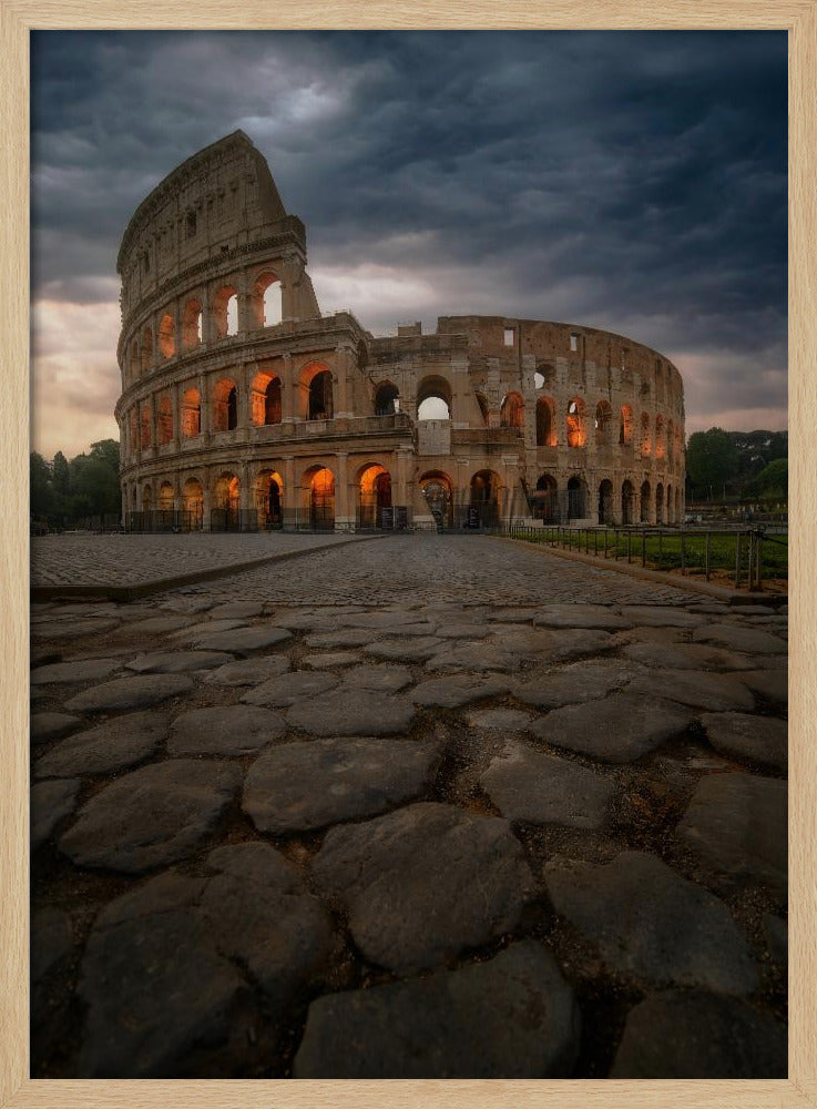 A low-angle view of the Roman Colosseum at dusk, with its arched windows glowing with warm orange light under a dramatic, stormy sky. An ancient cobblestone road leads towards the iconic amphitheater. Print