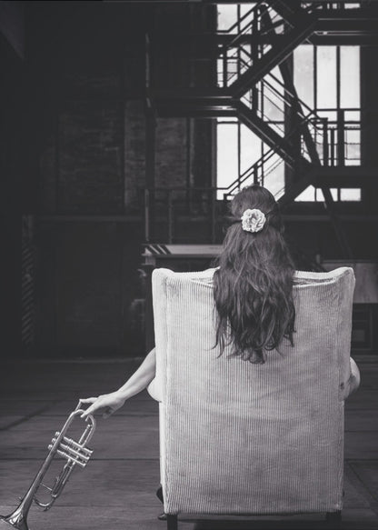 A black and white photograph of a woman seen from behind, sitting in a large, light-colored corduroy armchair. She has long, dark hair with a white flower pinned in it. Her left hand holds a trumpet, resting it on the dark floor of what appears to be an industrial warehouse with a metal staircase in the background. Wall Art