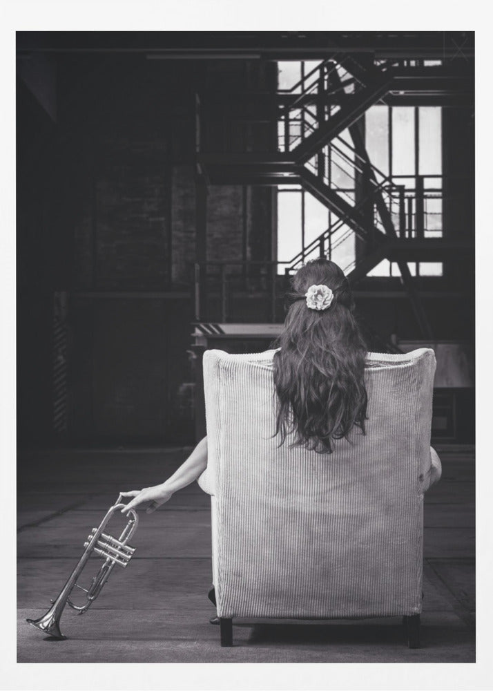 A black and white photograph of a woman seen from behind, sitting in a large, light-colored corduroy armchair. She has long, dark hair with a white flower pinned in it. Her left hand holds a trumpet, resting it on the dark floor of what appears to be an industrial warehouse with a metal staircase in the background. Wall Art