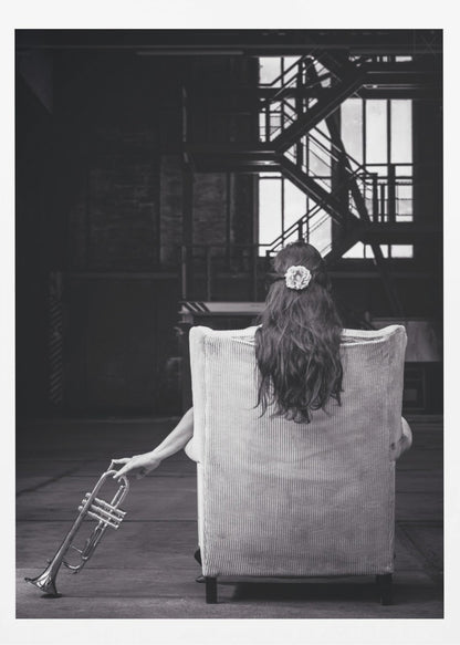 A black and white photograph of a woman seen from behind, sitting in a large, light-colored corduroy armchair. She has long, dark hair with a white flower pinned in it. Her left hand holds a trumpet, resting it on the dark floor of what appears to be an industrial warehouse with a metal staircase in the background. Wall Art