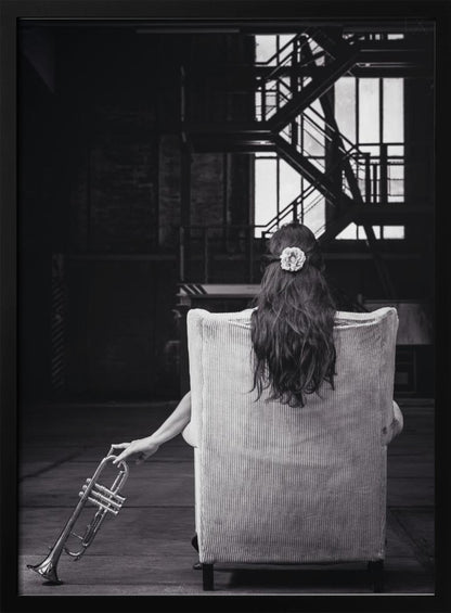 A black and white photograph of a woman seen from behind, sitting in a large, light-colored corduroy armchair. She has long, dark hair with a white flower pinned in it. Her left hand holds a trumpet, resting it on the dark floor of what appears to be an industrial warehouse with a metal staircase in the background. Wall Art
