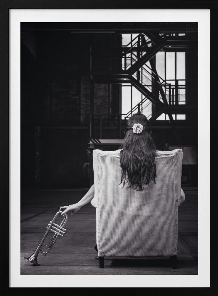 A black and white photograph of a woman seen from behind, sitting in a large, light-colored corduroy armchair. She has long, dark hair with a white flower pinned in it. Her left hand holds a trumpet, resting it on the dark floor of what appears to be an industrial warehouse with a metal staircase in the background. Wall Art