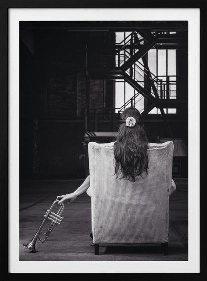 A black and white photograph of a woman seen from behind, sitting in a large, light-colored corduroy armchair. She has long, dark hair with a white flower pinned in it. Her left hand holds a trumpet, resting it on the dark floor of what appears to be an industrial warehouse with a metal staircase in the background. Wall Art
