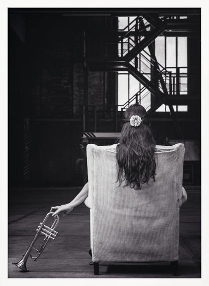 A black and white photograph of a woman seen from behind, sitting in a large, light-colored corduroy armchair. She has long, dark hair with a white flower pinned in it. Her left hand holds a trumpet, resting it on the dark floor of what appears to be an industrial warehouse with a metal staircase in the background. Wall Art
