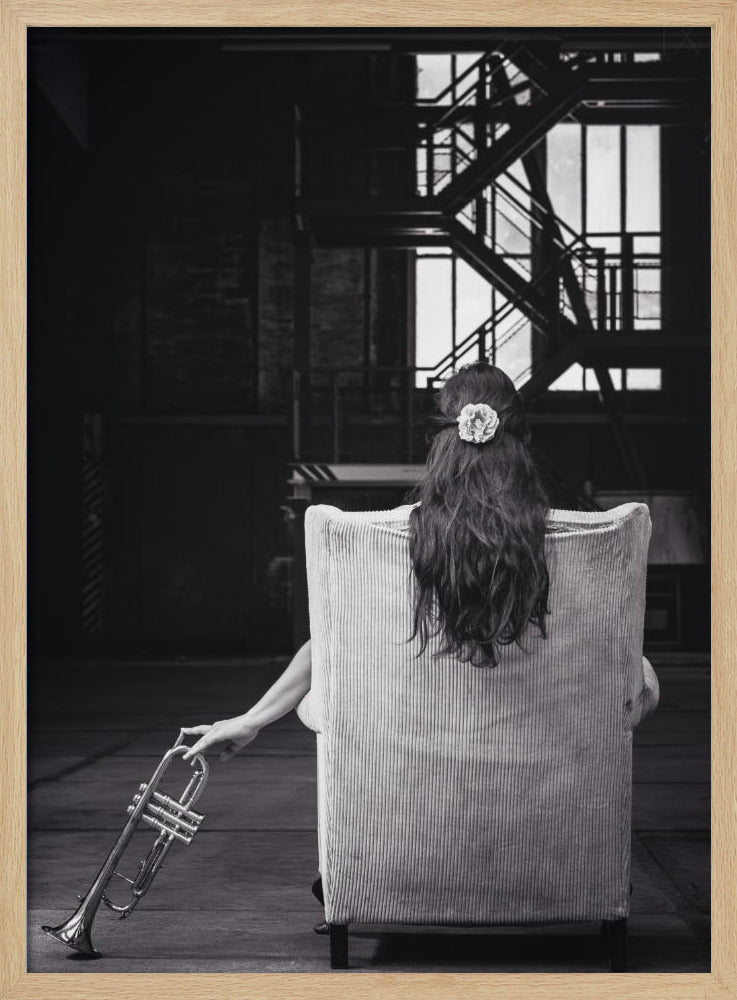 A black and white photograph of a woman seen from behind, sitting in a large, light-colored corduroy armchair. She has long, dark hair with a white flower pinned in it. Her left hand holds a trumpet, resting it on the dark floor of what appears to be an industrial warehouse with a metal staircase in the background. Wall Art