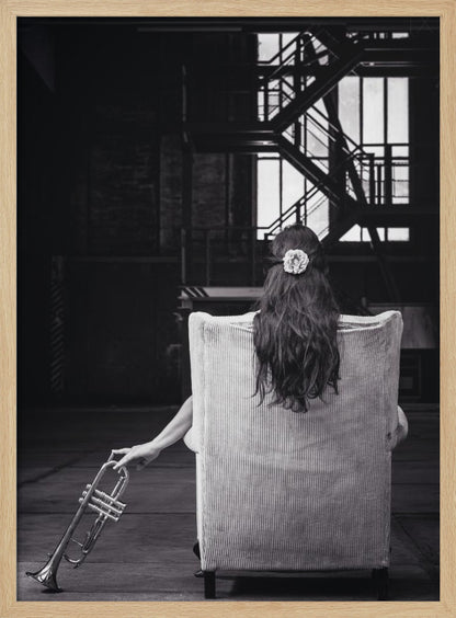 A black and white photograph of a woman seen from behind, sitting in a large, light-colored corduroy armchair. She has long, dark hair with a white flower pinned in it. Her left hand holds a trumpet, resting it on the dark floor of what appears to be an industrial warehouse with a metal staircase in the background. Wall Art