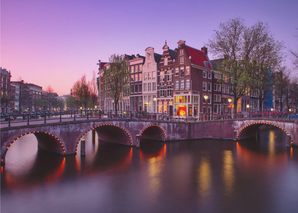 A scenic view of an Amsterdam canal at twilight, with historic brick buildings and illuminated arched bridges reflected in the calm water under a vibrant pink and purple sky. Poster