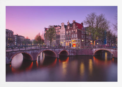 A scenic view of an Amsterdam canal at twilight, with historic brick buildings and illuminated arched bridges reflected in the calm water under a vibrant pink and purple sky. Poster