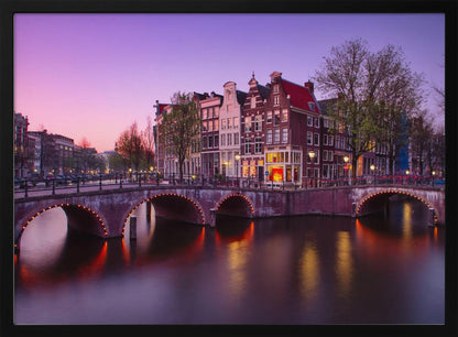 A scenic view of an Amsterdam canal at twilight, with historic brick buildings and illuminated arched bridges reflected in the calm water under a vibrant pink and purple sky. Poster