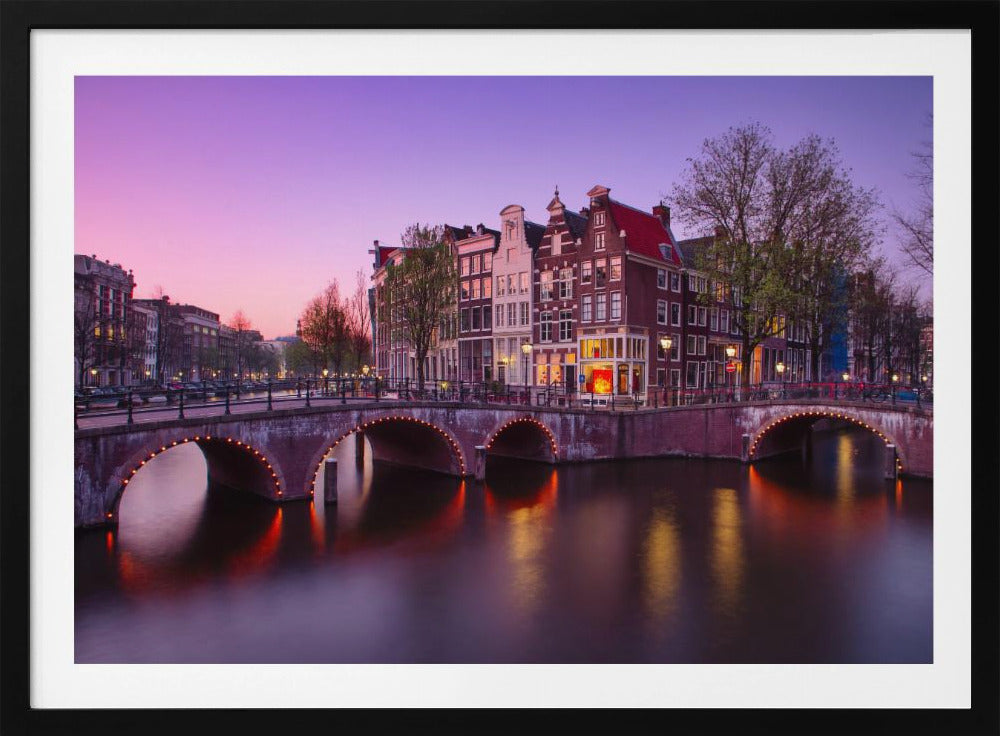 A scenic view of an Amsterdam canal at twilight, with historic brick buildings and illuminated arched bridges reflected in the calm water under a vibrant pink and purple sky. Poster