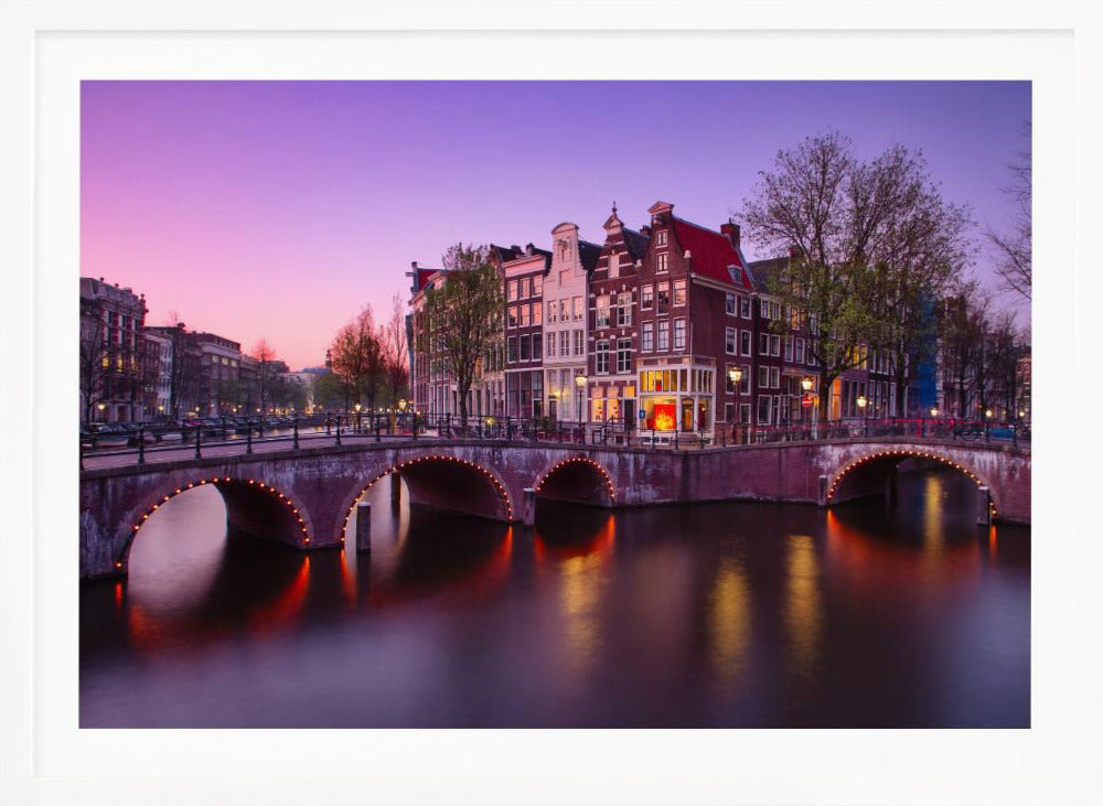 A scenic view of an Amsterdam canal at twilight, with historic brick buildings and illuminated arched bridges reflected in the calm water under a vibrant pink and purple sky. Poster