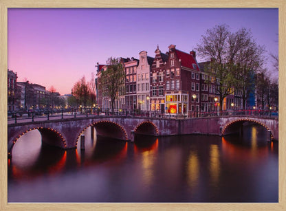 A scenic view of an Amsterdam canal at twilight, with historic brick buildings and illuminated arched bridges reflected in the calm water under a vibrant pink and purple sky. Poster