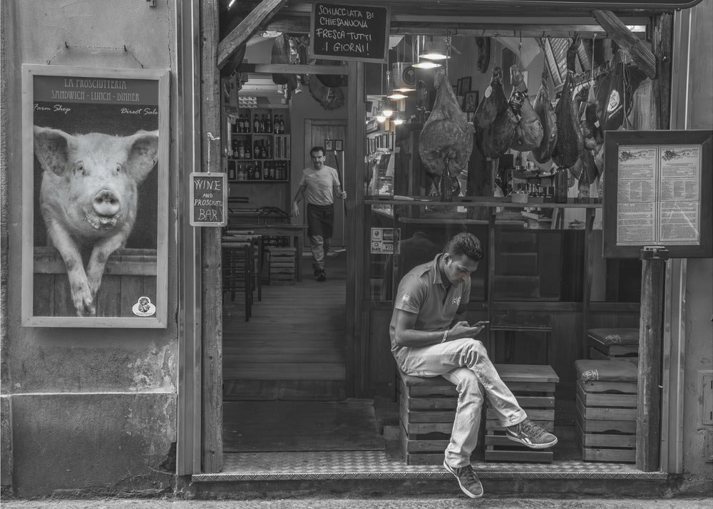 A framed, black and white street photograph of an Italian prosciutto shop. A young man sits on a wooden crate outside, looking at his phone, while cured hams hang inside the shop and a large, framed picture of a pig is on the exterior wall. Artwork