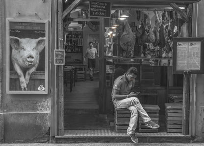 A framed, black and white street photograph of an Italian prosciutto shop. A young man sits on a wooden crate outside, looking at his phone, while cured hams hang inside the shop and a large, framed picture of a pig is on the exterior wall. Artwork
