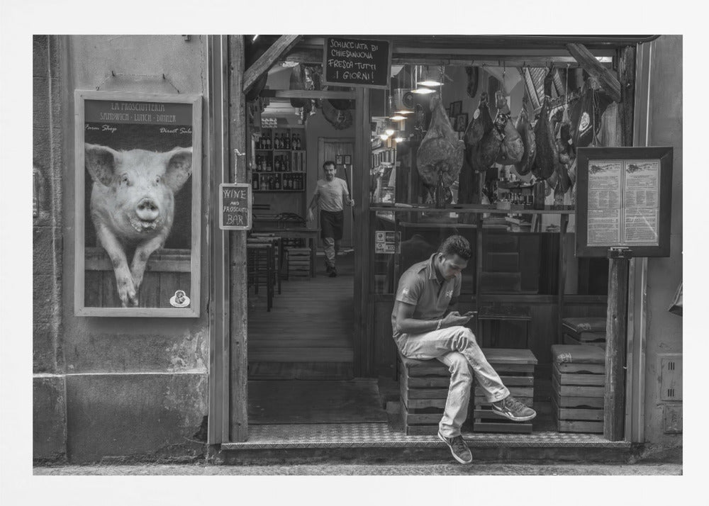 A framed, black and white street photograph of an Italian prosciutto shop. A young man sits on a wooden crate outside, looking at his phone, while cured hams hang inside the shop and a large, framed picture of a pig is on the exterior wall. Artwork