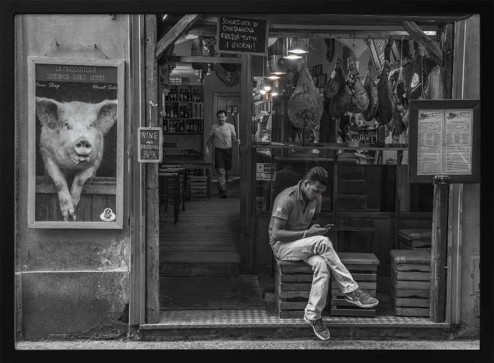 A framed, black and white street photograph of an Italian prosciutto shop. A young man sits on a wooden crate outside, looking at his phone, while cured hams hang inside the shop and a large, framed picture of a pig is on the exterior wall. Artwork