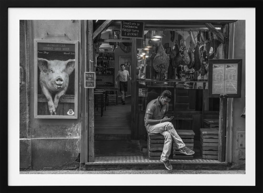 A framed, black and white street photograph of an Italian prosciutto shop. A young man sits on a wooden crate outside, looking at his phone, while cured hams hang inside the shop and a large, framed picture of a pig is on the exterior wall. Artwork