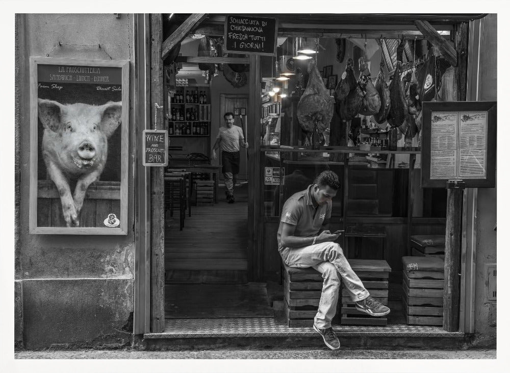 A framed, black and white street photograph of an Italian prosciutto shop. A young man sits on a wooden crate outside, looking at his phone, while cured hams hang inside the shop and a large, framed picture of a pig is on the exterior wall. Artwork