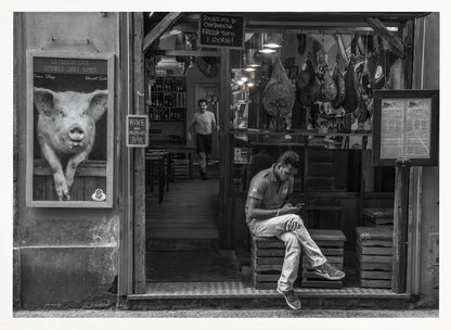A framed, black and white street photograph of an Italian prosciutto shop. A young man sits on a wooden crate outside, looking at his phone, while cured hams hang inside the shop and a large, framed picture of a pig is on the exterior wall. Artwork