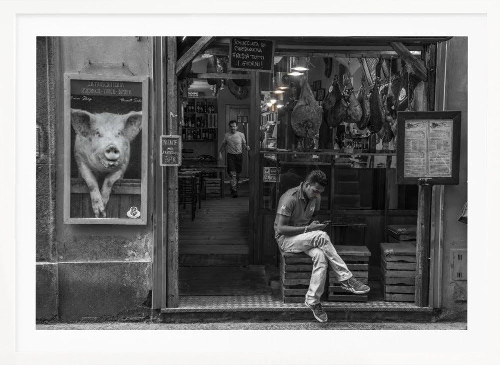 A framed, black and white street photograph of an Italian prosciutto shop. A young man sits on a wooden crate outside, looking at his phone, while cured hams hang inside the shop and a large, framed picture of a pig is on the exterior wall. Artwork