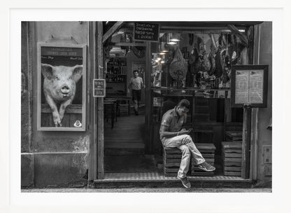 A framed, black and white street photograph of an Italian prosciutto shop. A young man sits on a wooden crate outside, looking at his phone, while cured hams hang inside the shop and a large, framed picture of a pig is on the exterior wall. Artwork