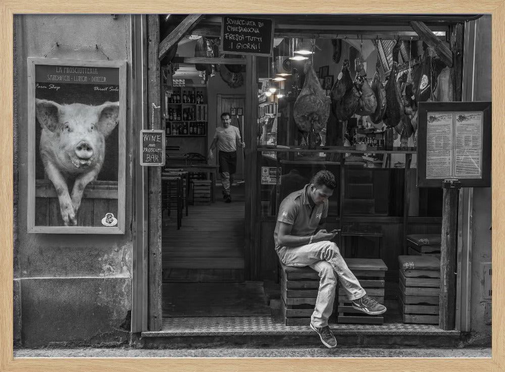 A framed, black and white street photograph of an Italian prosciutto shop. A young man sits on a wooden crate outside, looking at his phone, while cured hams hang inside the shop and a large, framed picture of a pig is on the exterior wall. Artwork