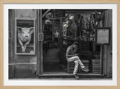 A framed, black and white street photograph of an Italian prosciutto shop. A young man sits on a wooden crate outside, looking at his phone, while cured hams hang inside the shop and a large, framed picture of a pig is on the exterior wall. Artwork