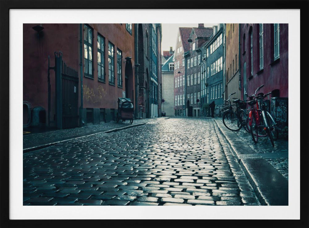 A low-angle shot of a wet, reflective cobblestone street in a narrow European alley. The street is lined with historic, colorful buildings in shades of red, orange, and blue under an overcast sky. Bicycles are parked along the sidewalk on the right, adding to the quiet, moody city scene. Poster