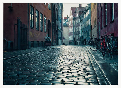 A low-angle shot of a wet, reflective cobblestone street in a narrow European alley. The street is lined with historic, colorful buildings in shades of red, orange, and blue under an overcast sky. Bicycles are parked along the sidewalk on the right, adding to the quiet, moody city scene. Poster