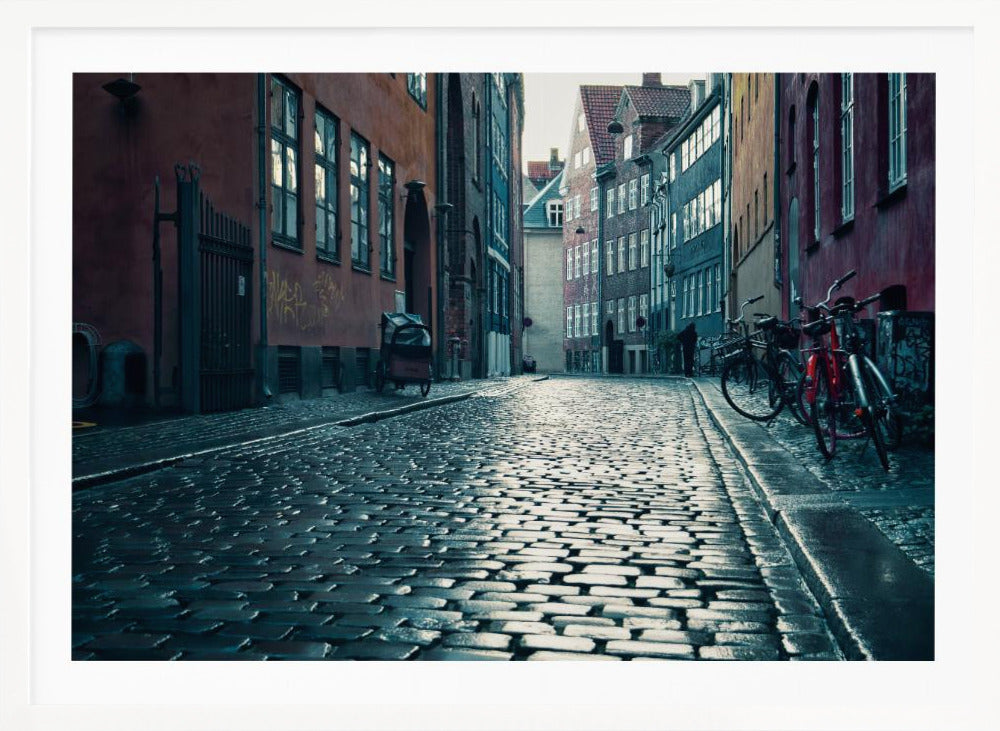A low-angle shot of a wet, reflective cobblestone street in a narrow European alley. The street is lined with historic, colorful buildings in shades of red, orange, and blue under an overcast sky. Bicycles are parked along the sidewalk on the right, adding to the quiet, moody city scene. Poster