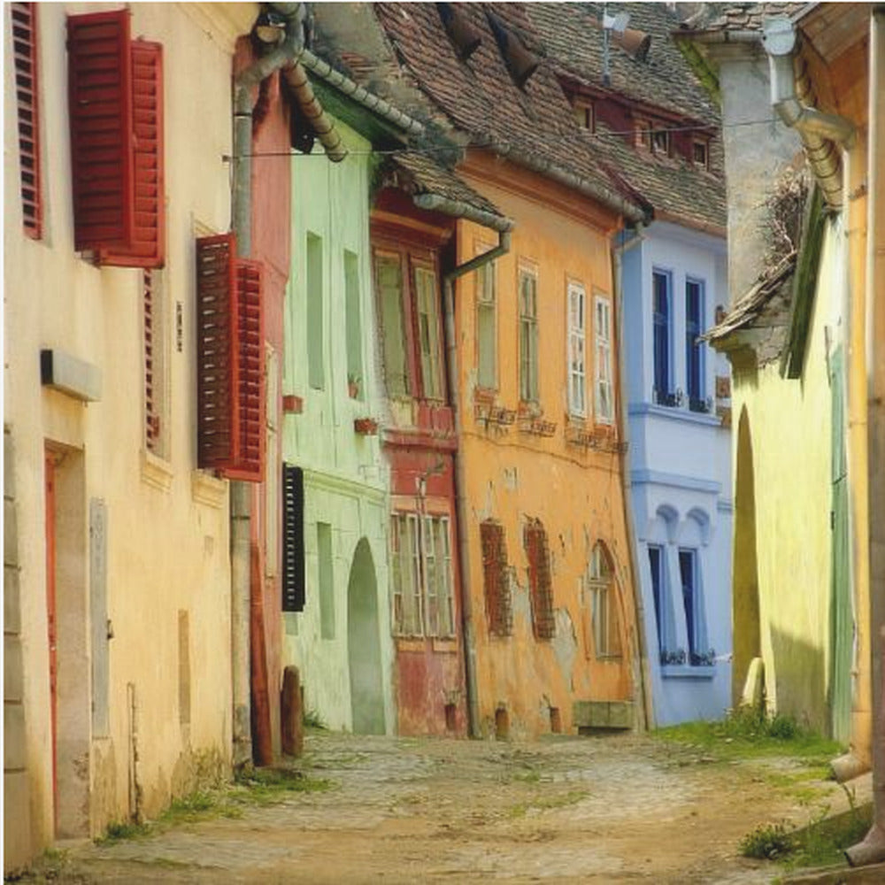 A narrow cobblestone street winds through a historic European town, flanked by colorful, weathered buildings in shades of cream, green, orange, and blue with red shutters, all under aged tile roofs and enclosed in a light wood frame. Artwork