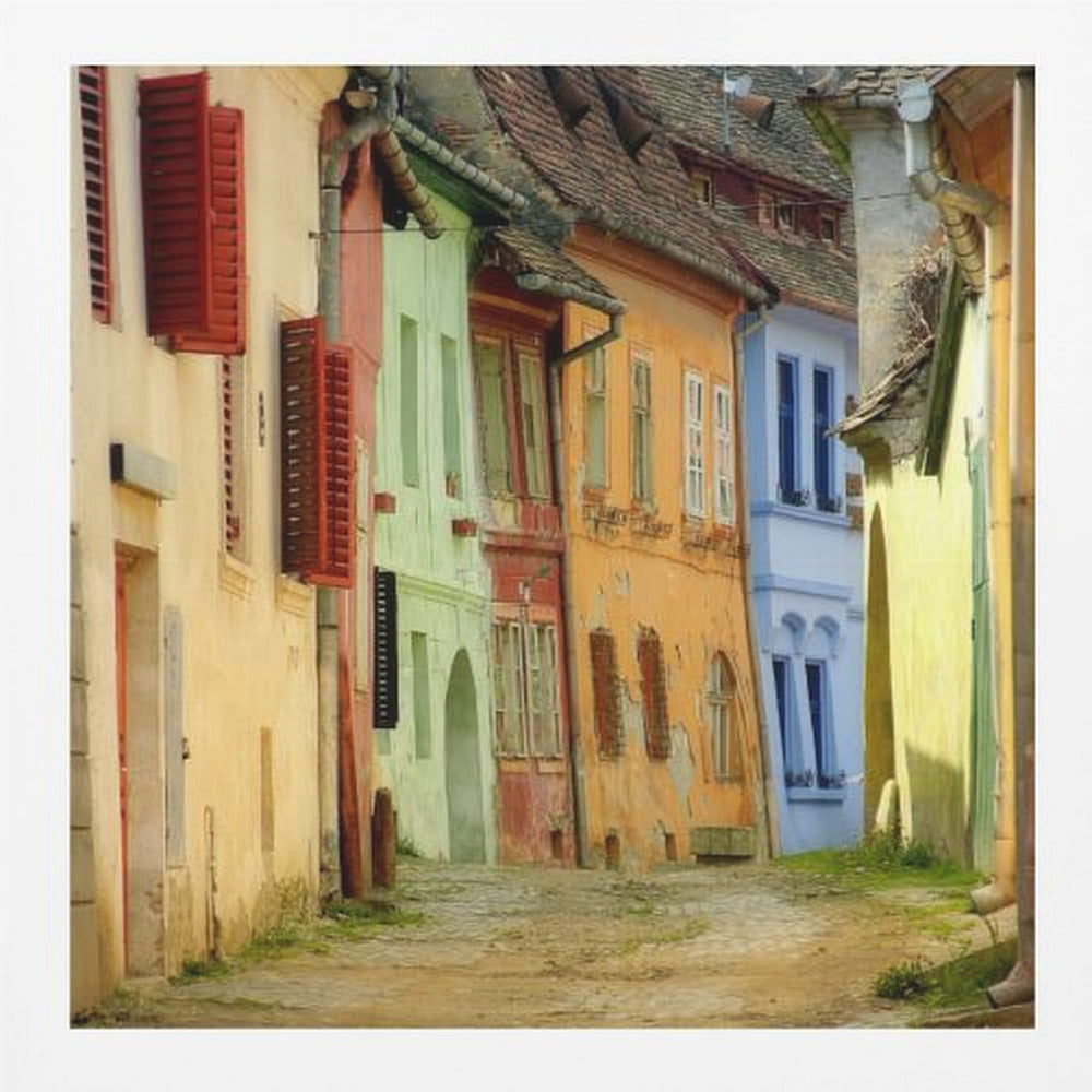 A narrow cobblestone street winds through a historic European town, flanked by colorful, weathered buildings in shades of cream, green, orange, and blue with red shutters, all under aged tile roofs and enclosed in a light wood frame. Artwork