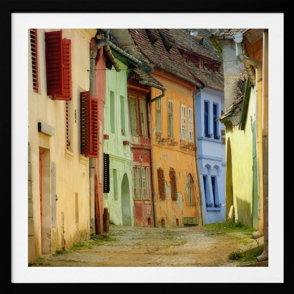 A narrow cobblestone street winds through a historic European town, flanked by colorful, weathered buildings in shades of cream, green, orange, and blue with red shutters, all under aged tile roofs and enclosed in a light wood frame. Artwork