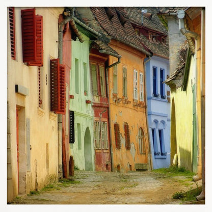 A narrow cobblestone street winds through a historic European town, flanked by colorful, weathered buildings in shades of cream, green, orange, and blue with red shutters, all under aged tile roofs and enclosed in a light wood frame. Artwork
