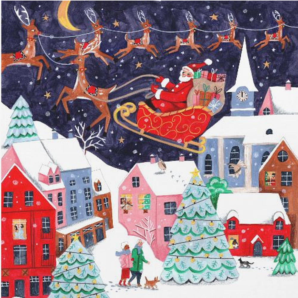 A festive illustration of Santa Claus flying in his sleigh pulled by reindeer over a snowy European-style village at night. The sky is a deep starry blue with a crescent moon, and the houses below are painted in vibrant reds, pinks, and blues, with warm light in the windows. People walk in the snow near decorated Christmas trees. Decor