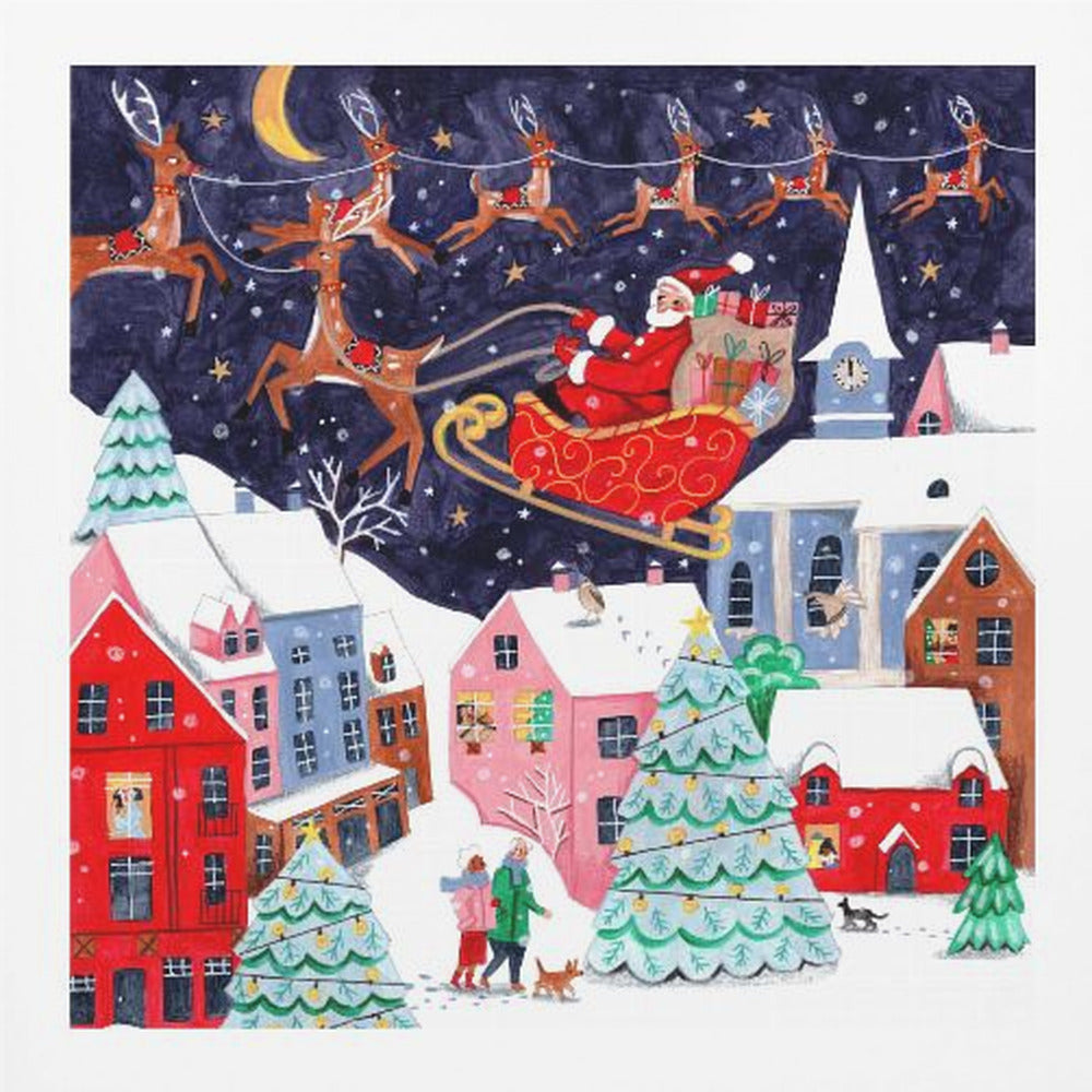A festive illustration of Santa Claus flying in his sleigh pulled by reindeer over a snowy European-style village at night. The sky is a deep starry blue with a crescent moon, and the houses below are painted in vibrant reds, pinks, and blues, with warm light in the windows. People walk in the snow near decorated Christmas trees. Decor
