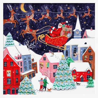 A festive illustration of Santa Claus flying in his sleigh pulled by reindeer over a snowy European-style village at night. The sky is a deep starry blue with a crescent moon, and the houses below are painted in vibrant reds, pinks, and blues, with warm light in the windows. People walk in the snow near decorated Christmas trees. Decor