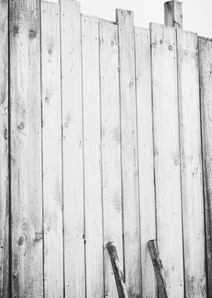 A black and white, low-angle photograph of a weathered wooden plank fence. The vertical planks are of varying heights, creating an uneven top line against a pale sky. The focus is on the texture of the wood, showing prominent grain, knots, and nail holes, all framed in black. Poster